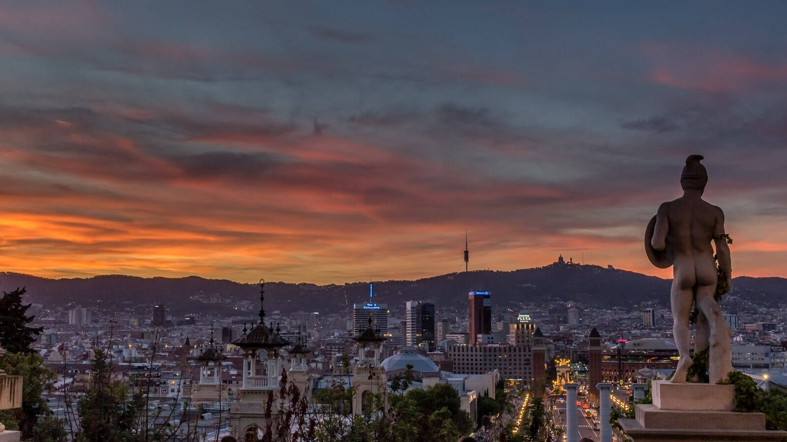 Une vue panoramique de Barcelone depuis Montjuïc, avec une statue en premier plan et la ville illuminée par les lumières du crépuscule ou de l'aube, sous un ciel orangé et nuageux.